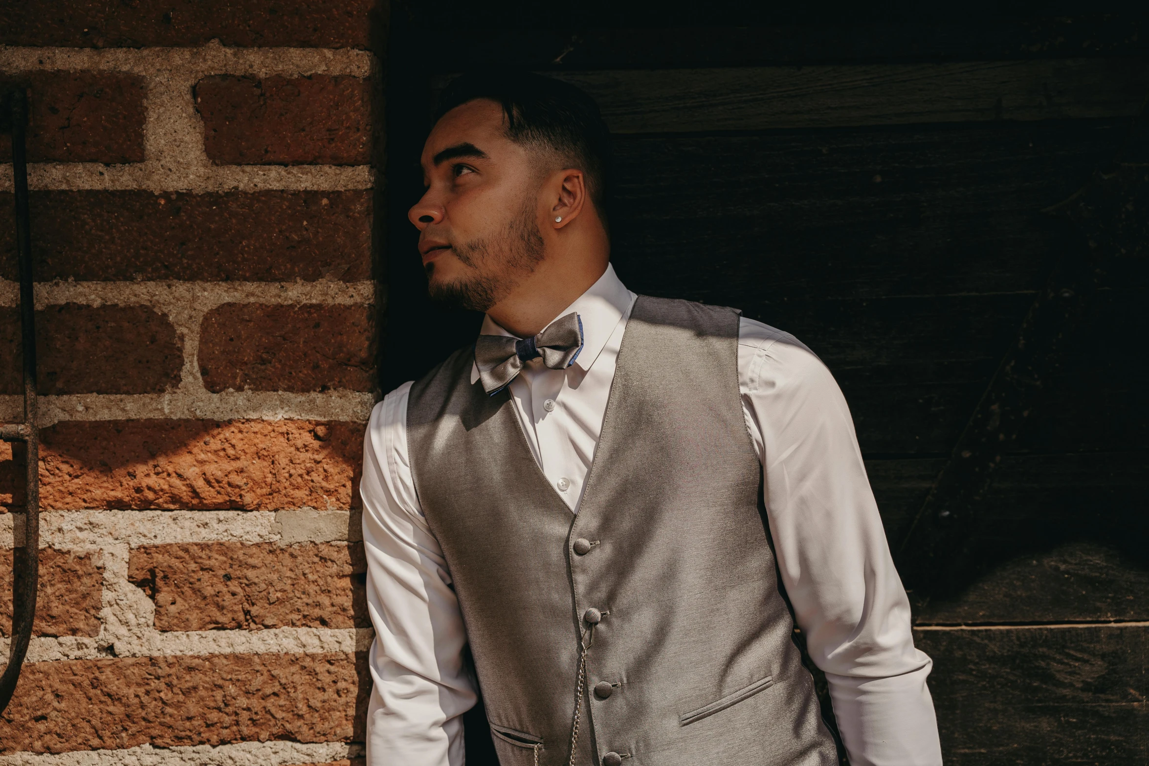 a man standing in front of a brick wall, platinum attire, bowtie, wearing a vest, profile image