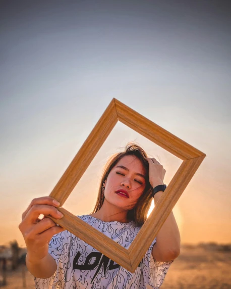 a woman holding a picture frame in front of her face, pexels contest winner, sunset lighting, non binary model, asian women, square face