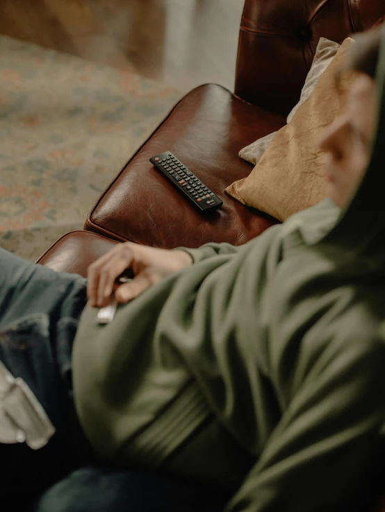 a man sitting on a couch holding a remote control, by Sebastian Spreng, trending on pexels, visual art, dusty rown bomber leather jacket, praying with tobacco, wearing a green sweater, low quality photo