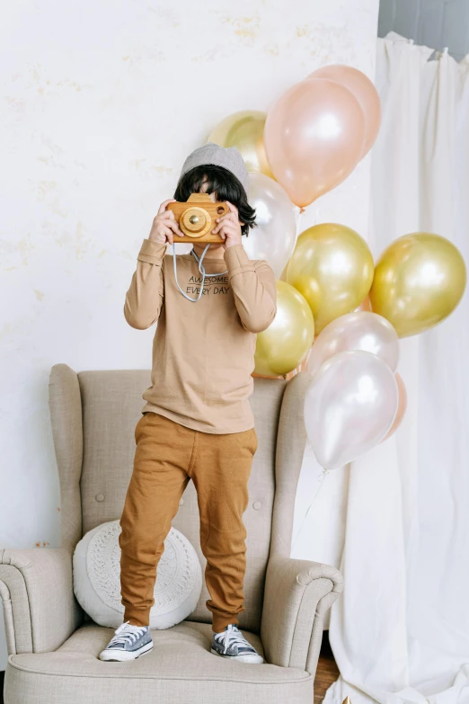 a boy standing in front of a chair holding a donut, a polaroid photo, party balloons, beige and gold tones, holding a dslr camera, long sleeves