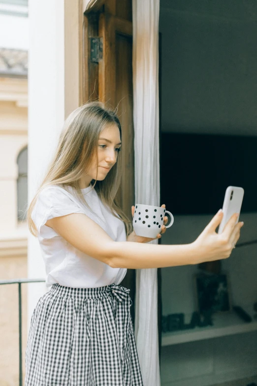 a woman standing on a balcony holding a cup of coffee, a picture, by Adam Marczyński, happening, mirror selfie, polka dot, a cute young woman, square