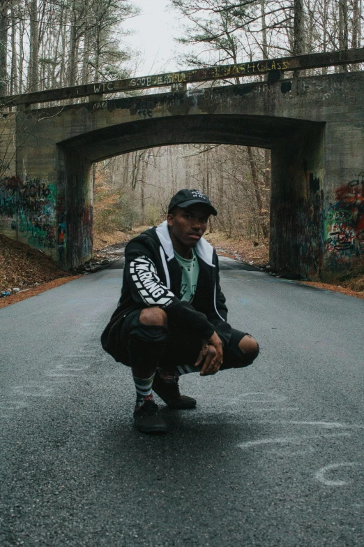 a man squatting down in the middle of a road, an album cover, unsplash, graffiti, in style of tyler mitchell, standing on a bridge, woods, rugged black clothes