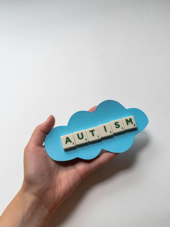 a person holding a scrabble that says autism, by Jessie Algie, aestheticism, nintendo clouds, diecut, high quality image, made of glazed