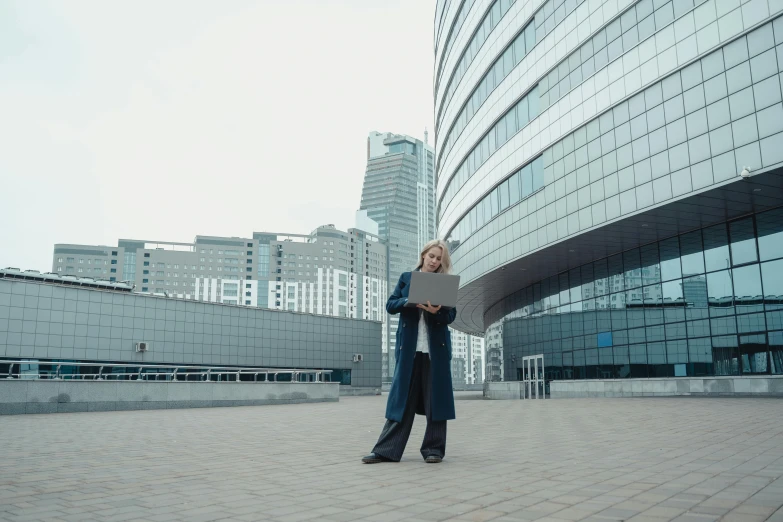 a woman holding a laptop standing in front of a building, pexels contest winner, modernism, russian city of the future, wearing a long coat, on the concrete ground, cosplay photo