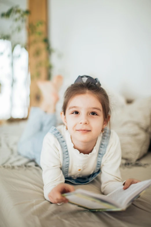 a little girl laying on a bed reading a book, pexels contest winner, lovingly looking at camera, confident relaxed pose, gif, kids