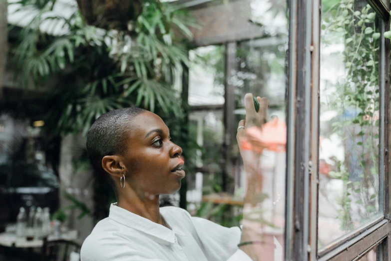 a woman in a white shirt looking out a window, by Lily Delissa Joseph, pexels contest winner, visual art, raining outside the cafe, wangechi mutu, lush surroundings, looking in mirror