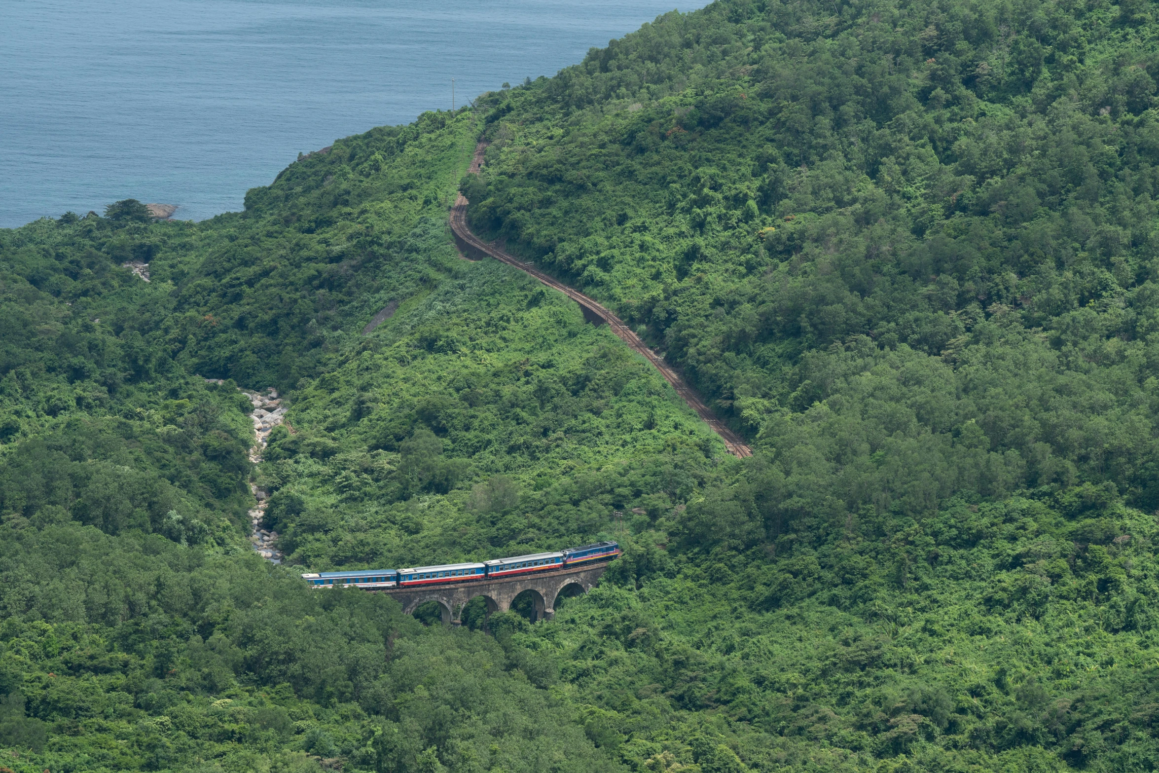 a train traveling over a bridge over a lush green hillside, near the sea, arik roper, indigo, on island