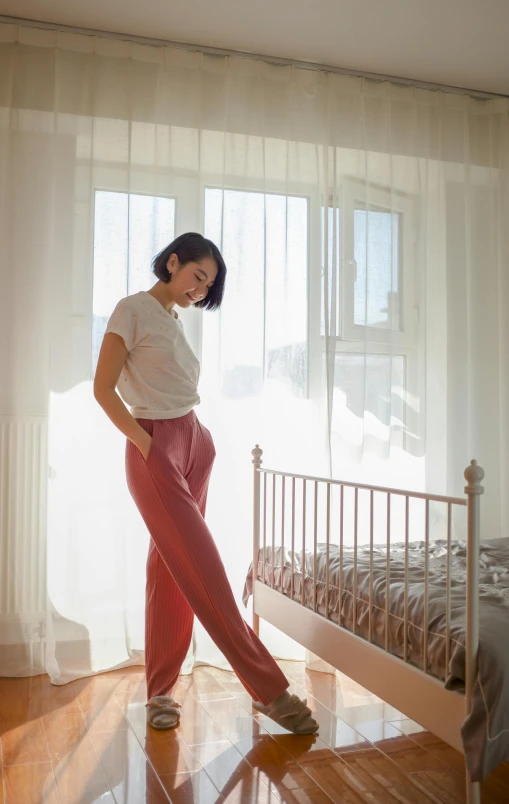 a woman standing in front of a bed in a bedroom, inspired by Judy Takács, pexels contest winner, arabesque, red shirt brown pants, kiko mizuhara, baggy pants, soft natural light