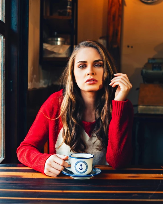 a woman sitting at a table with a cup of coffee, a portrait, inspired by Elsa Bleda, trending on unsplash, renaissance, red and blue garments, handsome girl, in a pub, frown fashion model