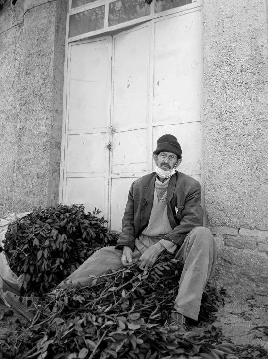 a black and white photo of a man sitting in front of a building, by Mahmoud Farshchian, covered in leaves, farmer, sage, 256435456k film