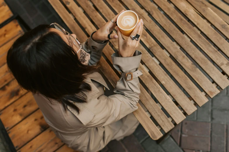 a woman sitting at a table with a cup of coffee, trending on pexels, light brown coat, sitting on bench, top down shot, manuka