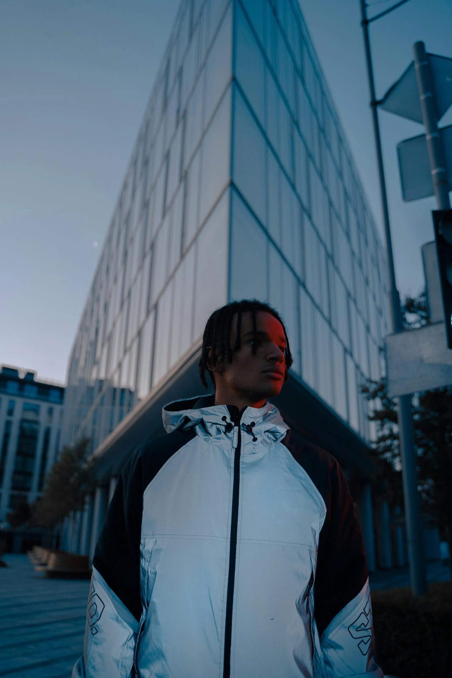 a man standing in front of a tall building, an album cover, unsplash, reflective puffer jacket, low quality footage, backlighted, wearing adidas clothing