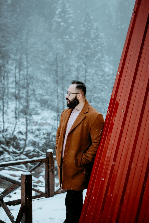 a man standing next to a red building in the snow, a picture, pexels contest winner, bearded beautiful man, on a bridge, brown colours, full body:: snow outside::