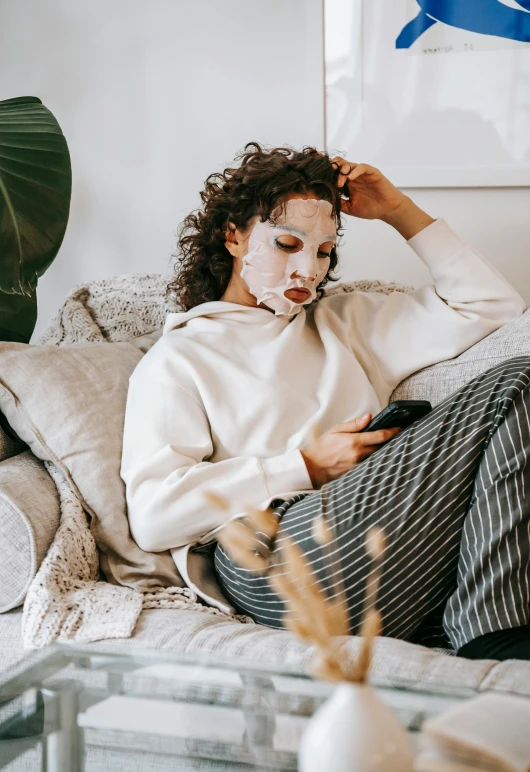 a woman sitting on a couch looking at her phone, trending on pexels, happening, face mask, sleepy fashion model face, white facepaint, messy bed