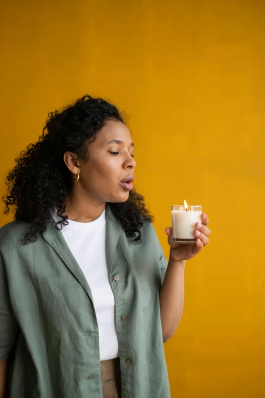 a woman holding a cup of coffee in front of a yellow wall, a portrait, by Nina Hamnett, trending on unsplash, renaissance, holding a candle, mixed-race woman, smelling good, waxy candles