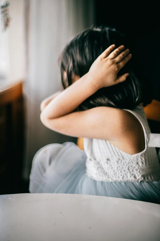 a little girl sitting at a table with her hands on her head, pexels, wearing a dress, haze over the shoulder shot, showing her shoulder from back, bedhead