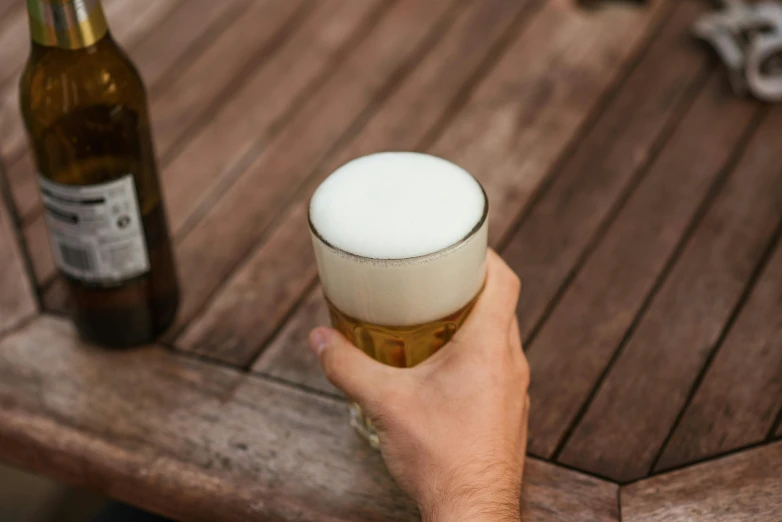 a close up of a person holding a glass of beer, white finish, on a wooden desk, manly, exterior shot