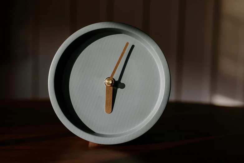 a clock sitting on top of a wooden table, a portrait, inspired by Lucio Fontana, unsplash, light grey blue and golden, matte finish, small, circle