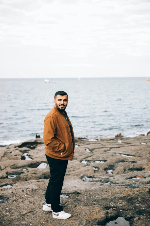 a man standing on top of a rock next to the ocean, a portrait of rahul kohli, he is wearing a brown sweater, in barcelona, profile image