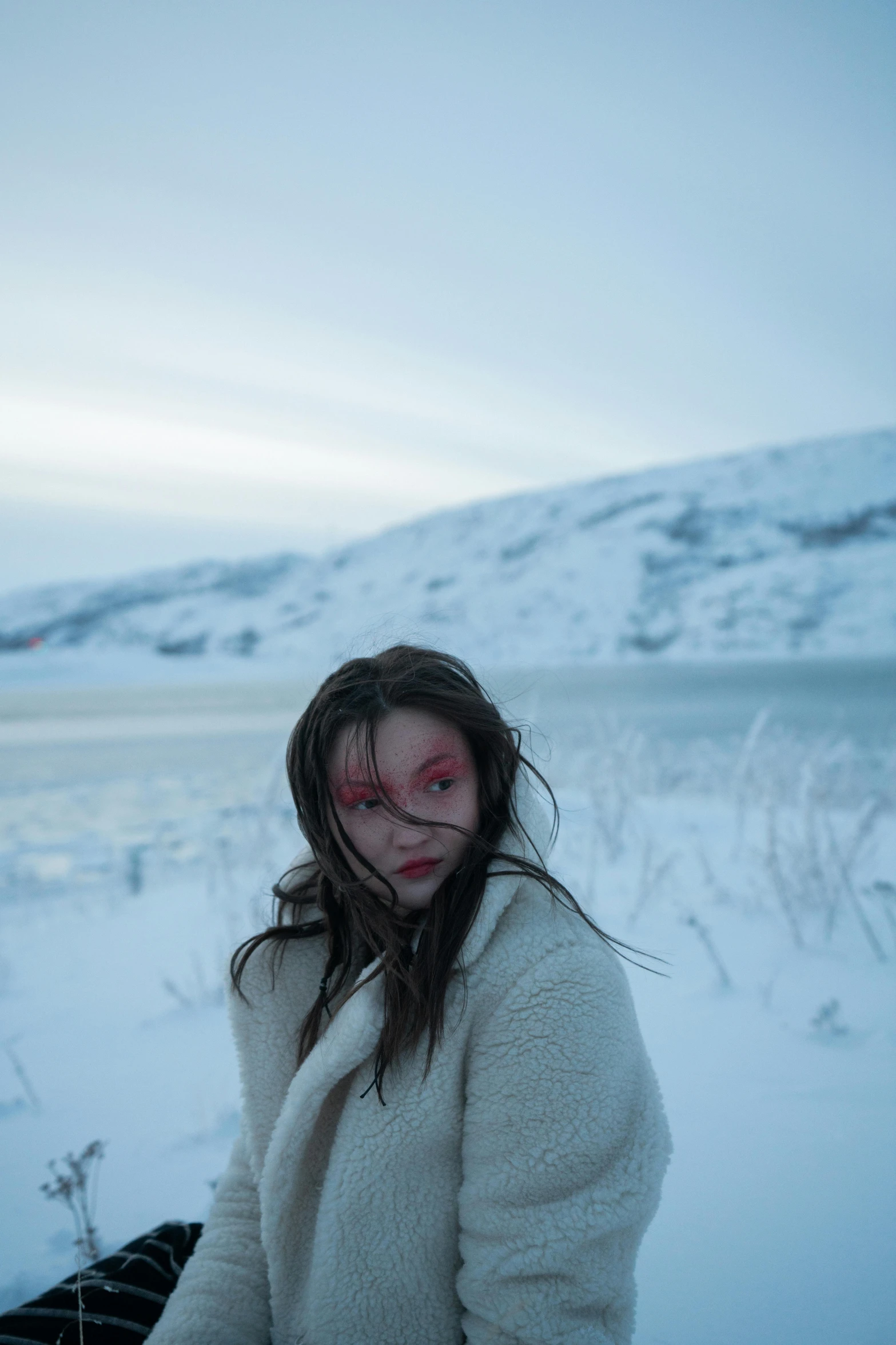 a woman sitting in the snow next to a body of water, an album cover, inspired by Elsa Bleda, pexels contest winner, with bloodshot eyes, iceland, actress, white facepaint