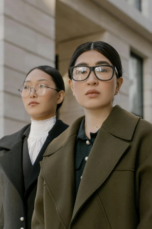 two women standing next to each other in front of a building, inspired by Wang Duo, trending on pexels, square rimmed glasses, looking serious, trench coat and suit, asian girl