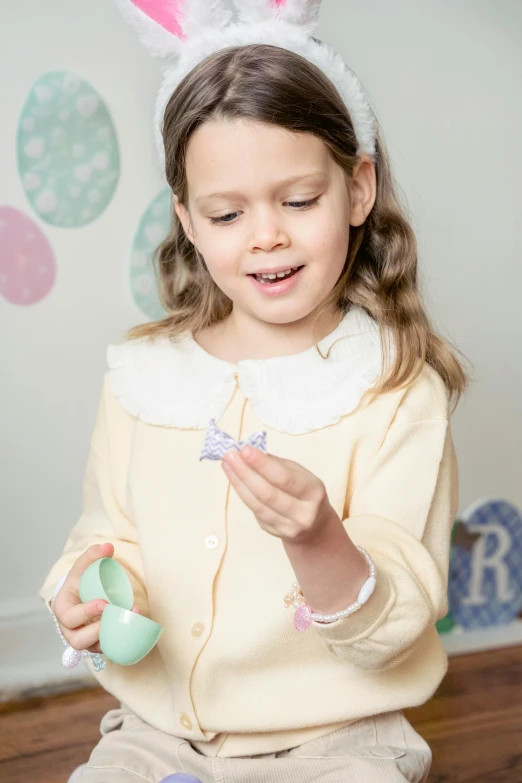 a little girl sitting on the floor holding an easter egg, inspired by Kate Greenaway, happening, product display photograph, tactile buttons and lights, pastel green, holding a bow