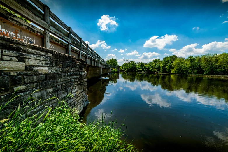 a bridge over a river next to a lush green field, by Andrew Domachowski, unsplash, visual art, blue sky, multiple stories, high details photo, thumbnail