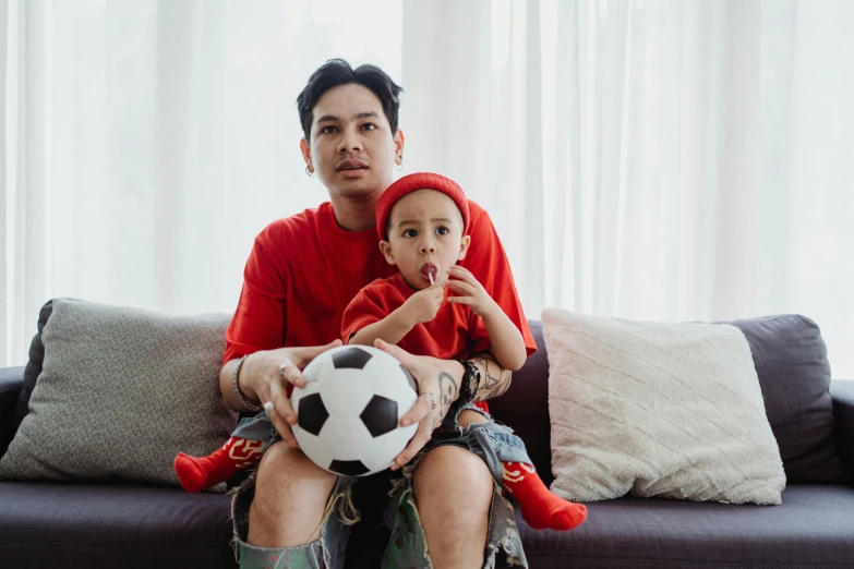 a man sitting on a couch holding a soccer ball, by Sebastian Spreng, pexels contest winner, an indonesian family portrait, bowater charlie and brom gerald, wearing red, lgbtq