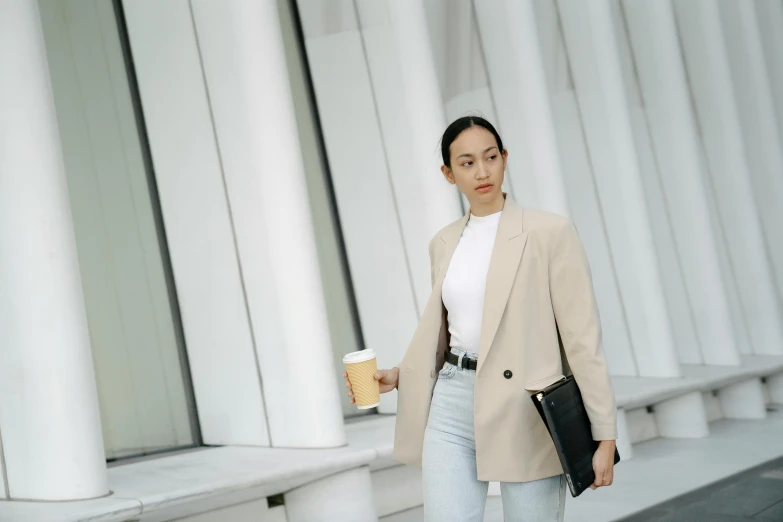 a woman walking down a sidewalk holding a cup of coffee, trending on pexels, minimalism, wearing a blazer, wearing off - white style, holding notebook, striking pose