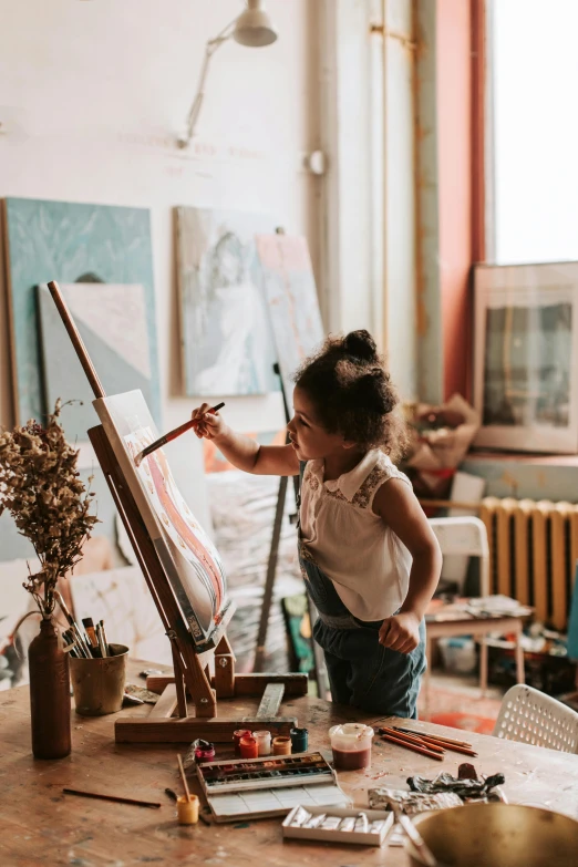 a little girl that is standing in front of a easel, pexels contest winner, painting a canvas, beautiful creative space behind, future activist, jpeg artefacts on canvas