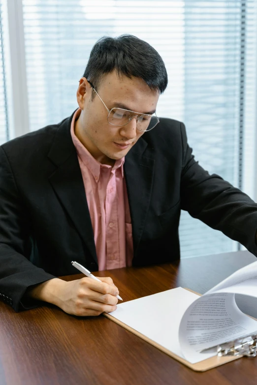 a man sitting at a desk writing on a piece of paper, asian male, wearing a suit and glasses, lgbtq, civil engineer