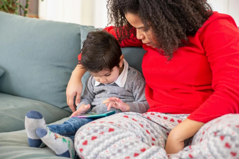 a woman and a child sitting on a couch, happening, red sweater and gray pants, looking at his phone, multicoloured, wearing pajamas