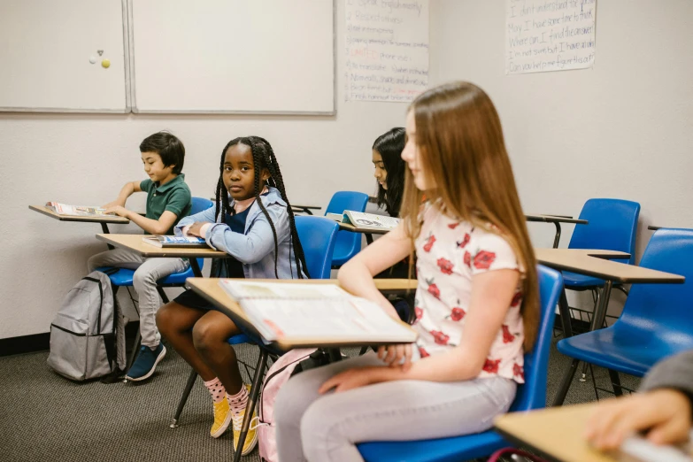 a group of children sitting at desks in a classroom, by Carey Morris, pexels, sitting on a chair, looking left, carson ellis, panoramic view of girl