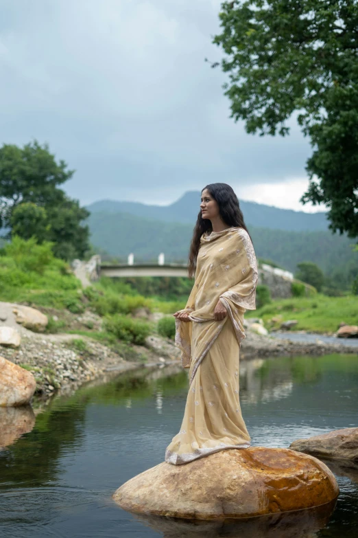 a woman standing on a rock in a river, inspired by Avigdor Arikha, renaissance, wearing sari, beige, in the countryside, roadside