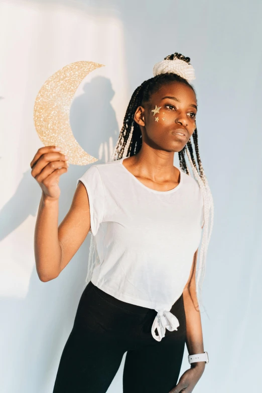 a woman standing in front of a wall holding a paper moon, by Dulah Marie Evans, dressed in a white t-shirt, white pearlescent, wearing a paper crown, young black woman