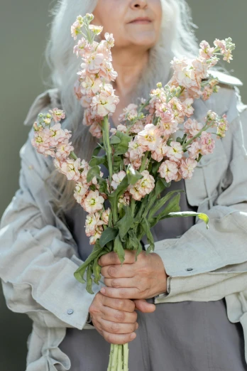 a woman holding a bunch of flowers in her hands, by Adriaen Hanneman, unsplash, wearing a light - pink suit, aged, wearing a pastel pink hoodie, elderly greek goddess