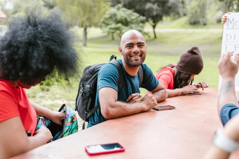 a group of people sitting around a wooden table, a portrait, pexels contest winner, happening, a man wearing a backpack, at the park on a beautiful day, brown skin man with a giant grin, sydney park