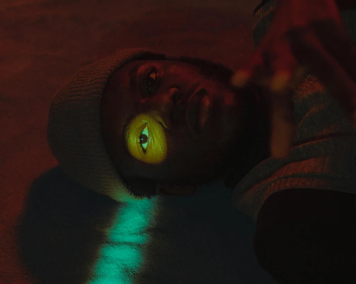 a man laying on the ground in the dark, by Liam Wong, his eyes glowing yellow, still from a music video, colored photo, ( ( dark skin ) )