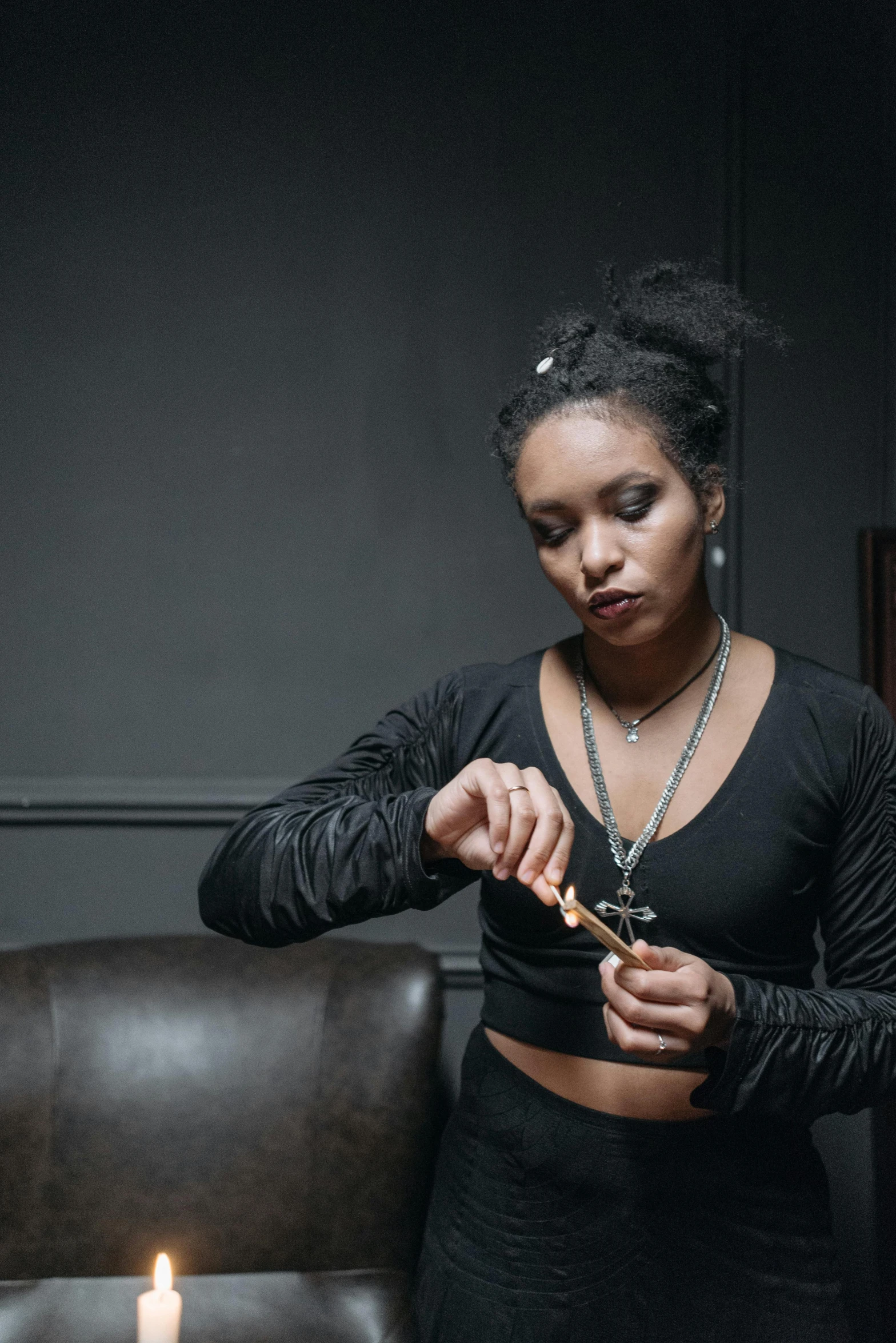 a woman lighting a candle in a dark room, an album cover, unsplash, leather jewelry, tessa thompson inspired, holding syringe, sitting on vintage leather sofa