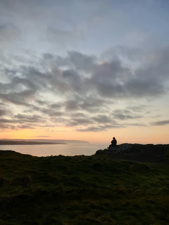 a person sitting on top of a grass covered hill, ocean in the distance, at dawn, shallan davar, phone photo