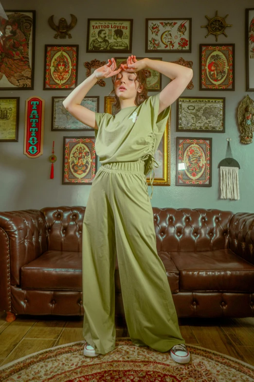 a woman standing in a living room next to a couch, a portrait, by Sven Erixson, unsplash, wearing atompunk jumpsuit, pastel green, wide leg hakama trousers, doing a sassy pose