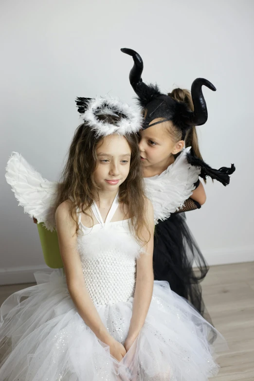 a couple of young girls sitting on top of a wooden floor, by Marie Angel, pexels, fantasy art, dream creature costumes, has black wings, two little horn on the head, with a white background