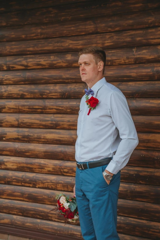 a man standing in front of a wooden wall, a colorized photo, by Pavel Fedotov, pexels contest winner, groom, button up shirt, flowers, cabin