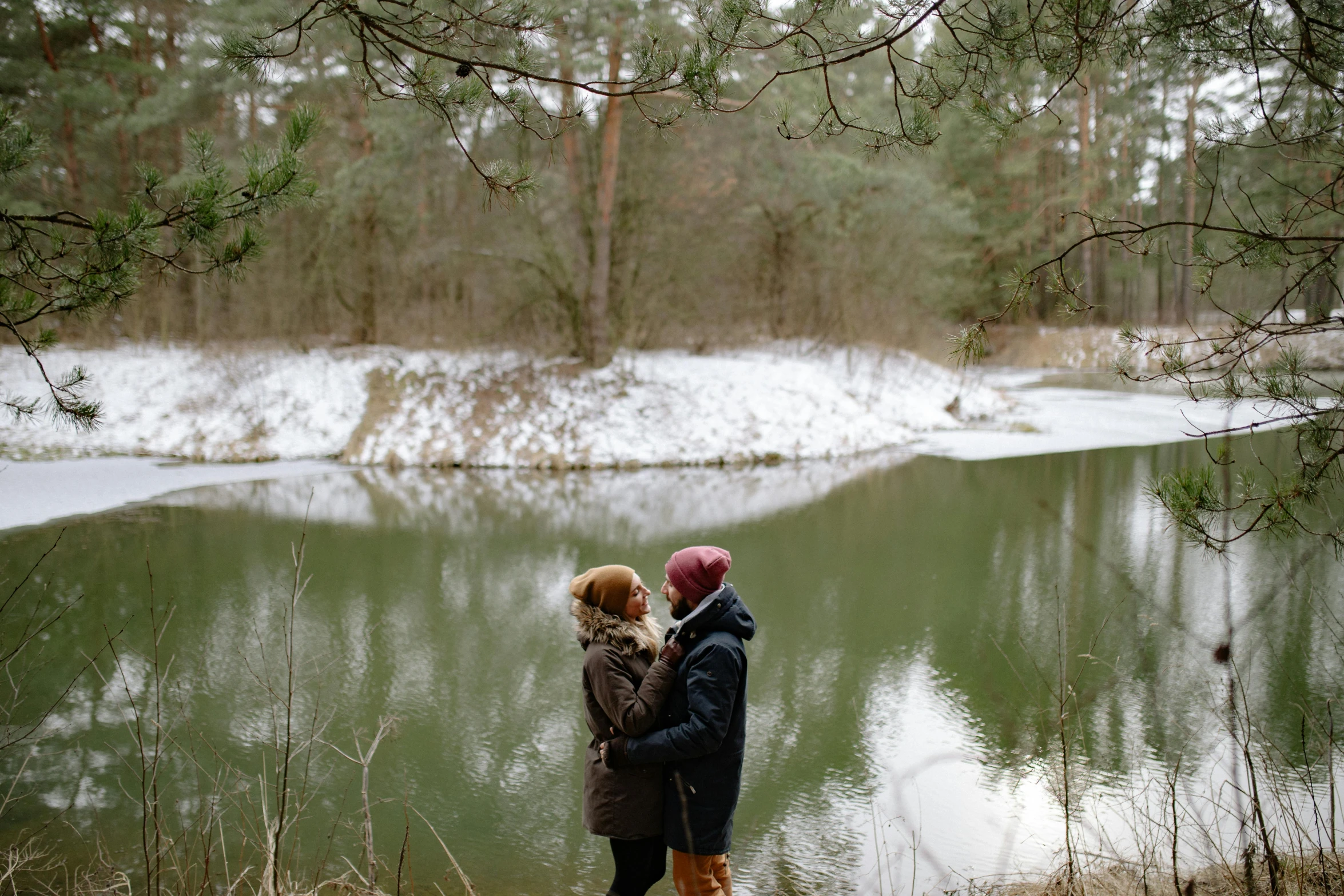 a couple of people standing next to a body of water, by Grytė Pintukaitė, pexels contest winner, winter forest, lesbian, cuddling, low quality photo