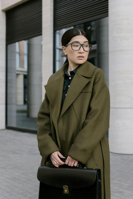 a woman standing in front of a building holding a briefcase, inspired by Feng Zhu, trending on pexels, wearing green jacket, half - frame square glasses, wearing long black winter coat, architect