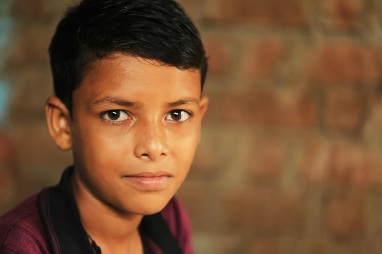 a young boy standing in front of a brick wall, ayan nag, in focus face with fine details, head and shoulders shot, looking straight into camera
