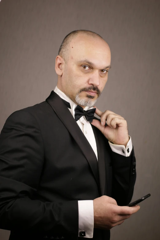 a man in a tuxedo holding a cell phone, an album cover, inspired by Cristache Gheorghiu, bald man, professional photo-n 3, headshot photograph, anton fadeev 8 k