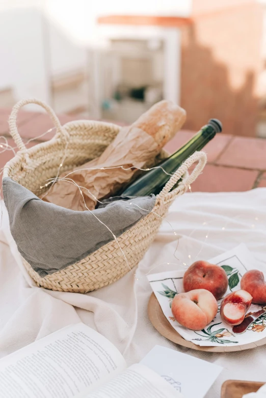 an open book sitting on top of a table next to a plate of fruit, fruits in a basket, holding wine bottle, bags on ground, linen