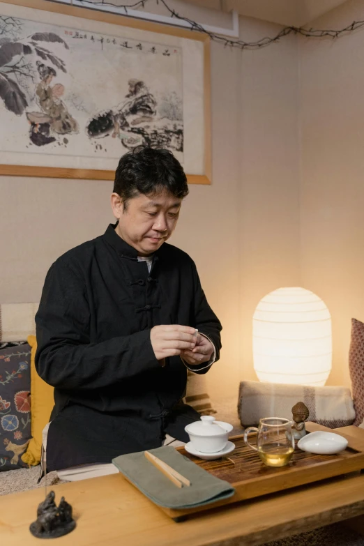 a man standing in a living room next to a table, inspired by Ma Lin, instagram, tea ceremony scene, partially cupping her hands, japanese collection product, evening lighting