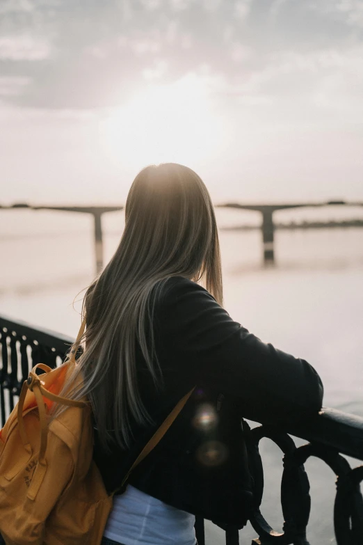 a woman standing on a bridge looking at the water, trending on pexels, looking off into the sunset, with a backpack, long hair girl, human staring blankly ahead